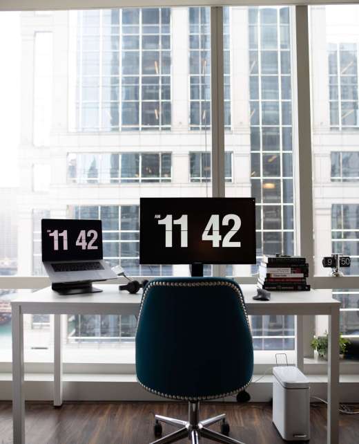 A vertical shot of a modern office desk with flat-screen monitors showing the time